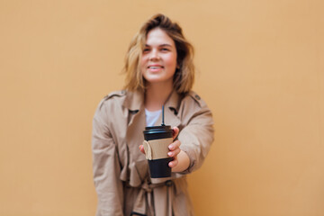 Fototapeta premium Young millennial woman with wild hair dressed in an autumn coat standing a cup of coffee to go near the wall.