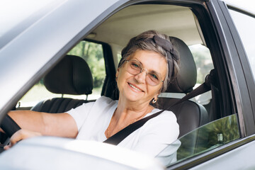 Joyful mature woman with glasses driving a car and looking at the camera through open window,...