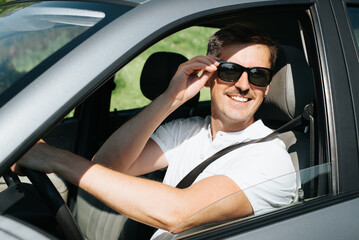 Fototapeta na wymiar Portrait of a joyful man in glasses driving a car and looking out the open window. Smiling handsome driver sitting behind the wheel of a car, outdoors. Happy road trip.