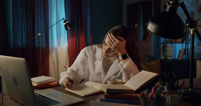 Tired young woman wearing medical uniform gown, female intern working at night with a computer. Doctor nurse using laptop and video call, writing notes, overtime at the workplace, headache.