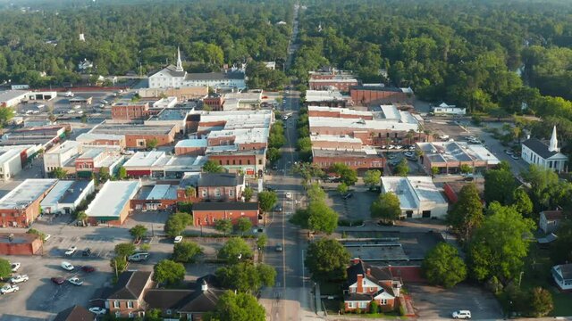 Aerial establishing shot of anytown America during golden hour. Businesses in historic town with large white church and steeple. Forest in distance.
