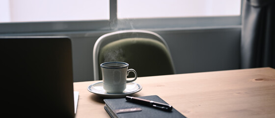 Fototapeta premium White cup of hot coffee, notebook and laptop computer on wooden table.