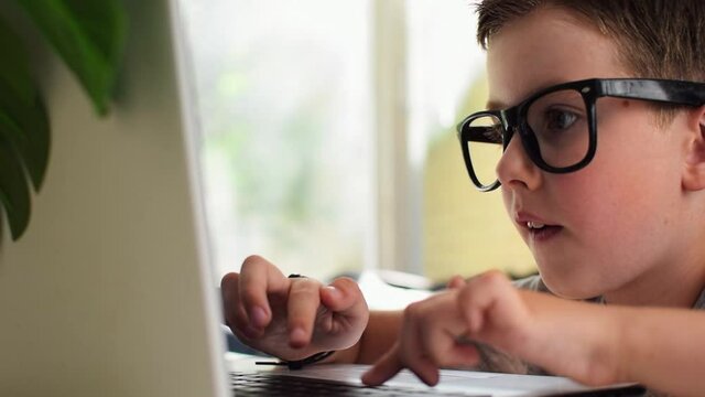 A smart boy in glasses looks at the laptop screen and learning programming at home. The child typing on keyboard. Kid play the game on computer. Side view, close-up. Kids online education concept.