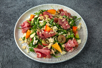 Salad with prosciutto, persimmon, almonds and dor blue cheese. Ready menu for the restaurant. Neutral gray blue textured background