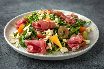 Salad with prosciutto, persimmon, almonds and dor blue cheese. Ready menu for the restaurant. Neutral gray blue textured background