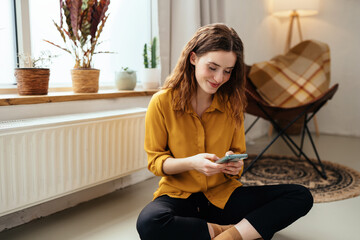 Young woman sitting in front of a radiator with her mobile phone - obrazy, fototapety, plakaty