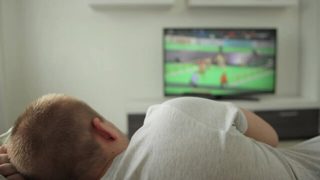 Man fan is lying on sofa in room at home and watching sports event on TV. Summer olimpic game competition. Athletics running 2021. Rooter buff watches broadcast on plasma in modern interior. Tokyo
