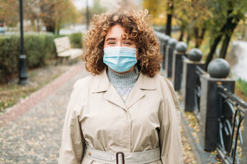 Pandemic outdoors. Covid prevention. New normal. Cheerful happy plus size woman with curly hair in medical face mask wearing outerwear in autumn park street.