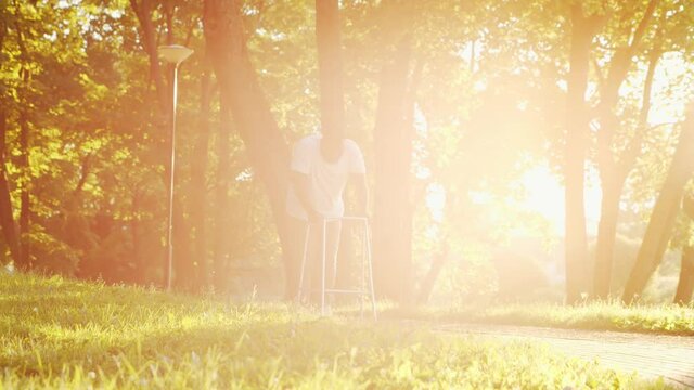 Disabled African-American man is walking with a walker. Handicapped patient in the park. Assistance, rehabilitation and health care. Golden hour sunset.