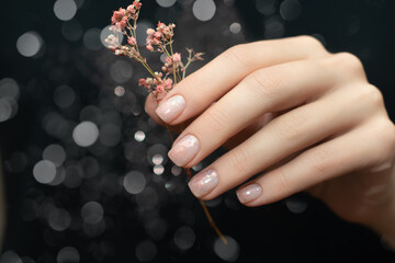Female hand with beige nail design. Glitter beige nail polish manicure. Woman hand hold pink flower on black background.