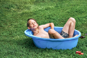 Relaxed boy lies in plastic basin on green lawn grass