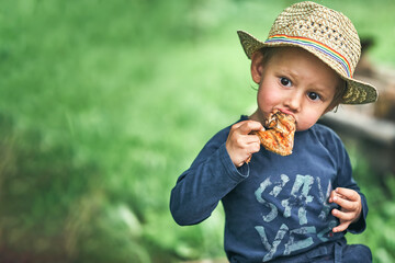 Boy in straw hat eats grilled chicken wing in green yard