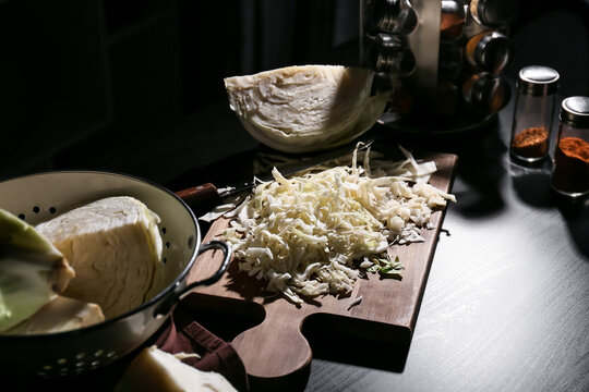 Freshly shredded cabbage on wooden cutting board with whole cabbage head and colander, kitchen preparation scene, a large half-head of cabbage rests nearby in a colander