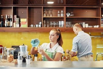 Serious young woman working in coffeeshop or restaurant, she is wiping glass jars when getting ready for opening in the morning