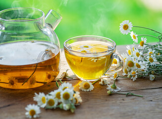 Herbal chamomile tea and chamomile flowers near teapot and tea glass on wooden table. Countryside background.
