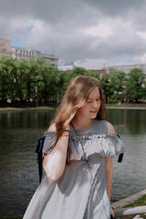 Smiling young woman in dress standing alone among street, leaning on fence in front of pond. looking at camera. Having fun outside. Urban style and street fashion. Sumeer casual outfit