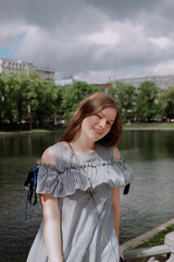 Smiling young woman in dress standing alone among street, leaning on fence in front of pond. looking at camera. Having fun outside. Urban style and street fashion. Sumeer casual outfit
