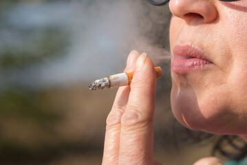 Middle age woman smoking cigarette outdoors with sun on her face. Closeup woman with cigarette in...