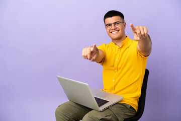 Young man sitting on a chair with laptop points finger at you while smiling