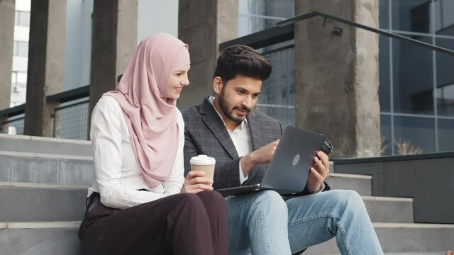 Woman in hijab and man in suit sitting on stairs near office building and discussing common project. Arabian partners using modern laptop for work outdoors. Two arabian colleagues sitting on stairs