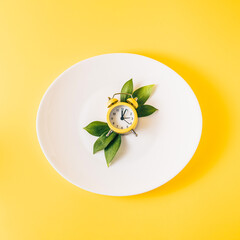 Clock with green leaves on white plate setup on pastel yellow background. Minimal time concept. Flat lay.