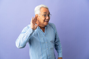 Middle age Brazilian man isolated on purple background listening to something by putting hand on the ear