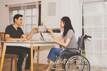 Fototapeta premium Disabled beautiful woman sitting on a wheelchair working with the male boyfriend in the home office, helping to deliver papers to the girlfriend who must help each other on the desk with understanding