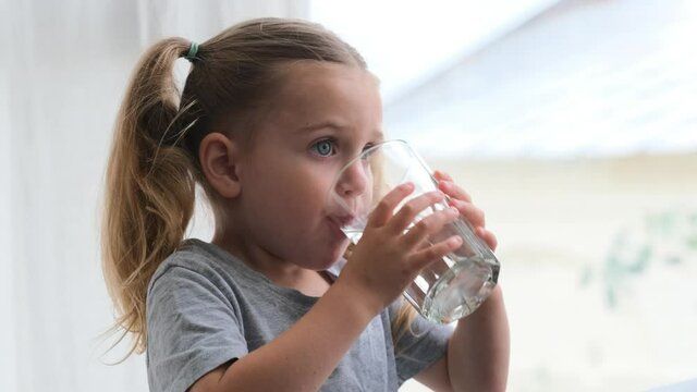 Little blonde girl in white dress drinks water from a glass indoors. Cute child is drinking a cup of water