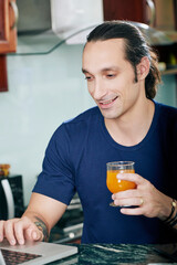 Portrait of smiling handsome man drinking fruit juice and checking e-mails on laptop when sitting at kitchen counter