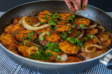 Woman's hand sprinkles parsley over sweet and sour chicken patties in a pan, on a plaid towel. Chicken cutlets cooked with Asian flavored sauce and caramelized onions. Chinese style dish, side view.