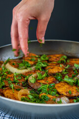 Woman's hand sprinkles parsley over sweet and sour chicken patties in a pan, on a plaid towel. Chicken cutlets cooked with Asian flavored sauce and caramelized onions. Chinese style dish, side view.