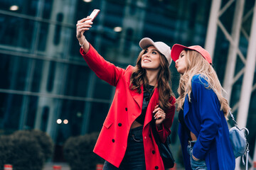 Two joyful cheerful girls taking a selfie while standing together at street near the mall