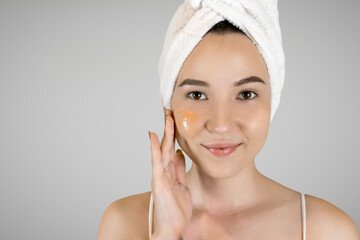 Beauty portrait of smiling woman in towel on head applying yellow nourishing mask on face on gray isolated background. Skin care concept. 