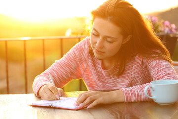 Woman writing on agenda at sunset in a balcony