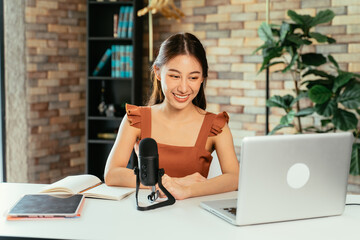 Portrait of cheerful young Asian female blogger using mic and laptop with digital tablet to...
