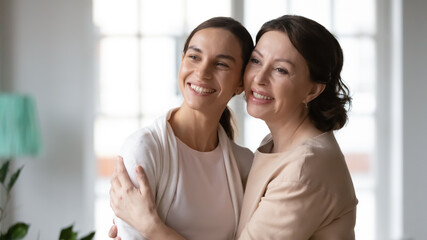 Smiling young adult girl and older Caucasian mom hug cuddle look in distance dream think of happy future together. Happy grownup daughter and middle-aged mother embrace show love and care.