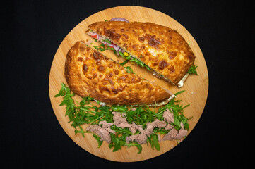 Tuna sandwich, cheese sauce, tomatoes, red onions, corn and arugula salad on a wooden plate on a black background.