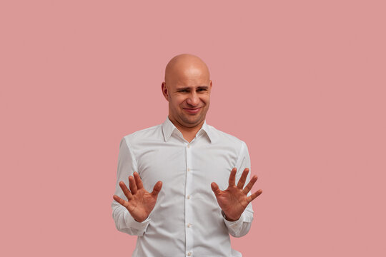 It is disgusting. Dissatisfied bald guy with bristle wears white shirt frowns face in dislike, makes refusal gesture, sees something abominable loathsome. Isolated over pink background.