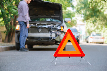 Red triangle warning sign of car accident on road. Triangle in front of wrecked car and driver man. Injured person man in accident looks under the hood of wrecked car after an accident.