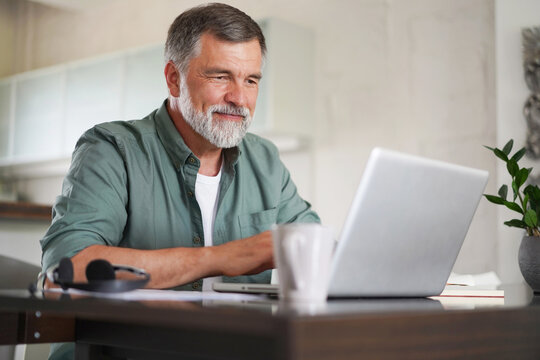 Handsome mature man in casual suit sitting at the table in home office and working at laptop