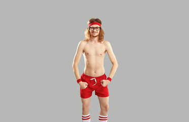 Funny topless skinny man in glasses, red retro sweatband and shorts standing isolated on gray...
