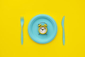 Intermittent fasting, lunch time, diet. Alarm clock, plate and cutlery. Yellow and blue colors.