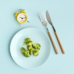 Intermittent fasting, diet, lunch time concept. Yellow alarm clock, broccoli in a plate and cutlery