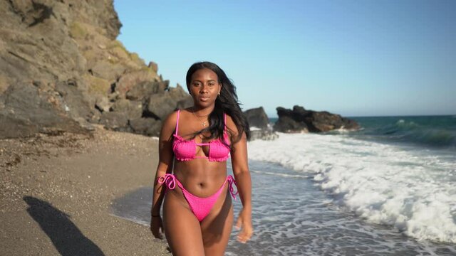 african american woman walking along the shore of the beach in a bikini