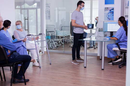 Nusre with visor against coronavirus and young patient talking in reception area, checking appointment informations. Senior disabled woman with walking frame discussing with assistant in waiting room.