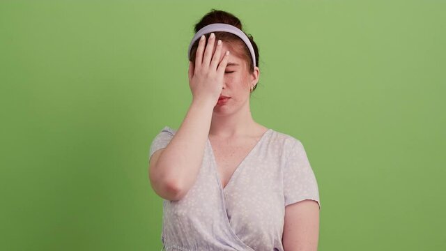 An upset redhead girl in a dress and with a rim on her head making a facepalm gesture in a green studio