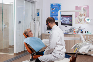 Dentist in dental clinic saying diagnostic to senior woman wearing lab coat. Medical teeth care taker talking with patient sitting on chair during consultation.