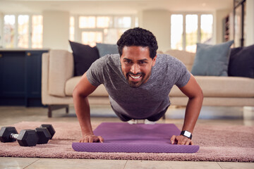 Fototapeta premium Man In Fitness Clothing At Home In Lounge Doing Press Ups And Exercising With Hand Weights