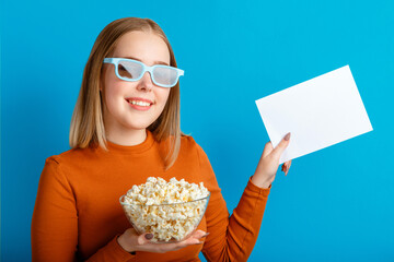 Emotional portrait of young woman in cinema glasses. Smiling teenager girl movie viewer in glasses holding popcorn and emty white blanc card for mock up copy space isolated over blue color background.