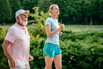 Side view of active beard senior man and woman physiotherapist running outside in green nature. Elder man and his adult sportive daughter exercising in the summer park, outdoor. Different generations.